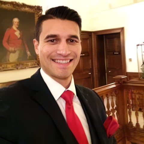 Man in black suit and red tie smiling at camera in an elegant formal interior with portrait and wooden doors