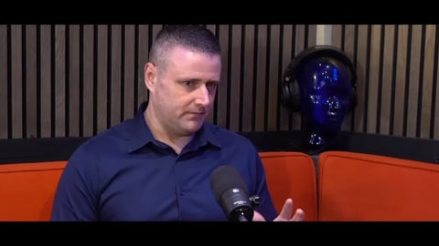 Man in navy blue shirt sitting on orange couch during podcast or interview with microphone
