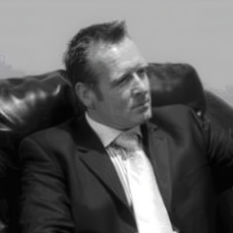 Black and white photograph of a man in a dark suit and white dress shirt, reclining on a leather chair with a contemplative expression