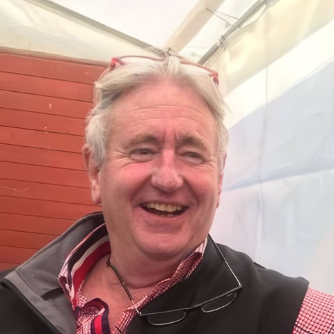 Smiling older man with gray hair wearing a black jacket and red checkered shirt, photographed outdoors with red and white structures in background