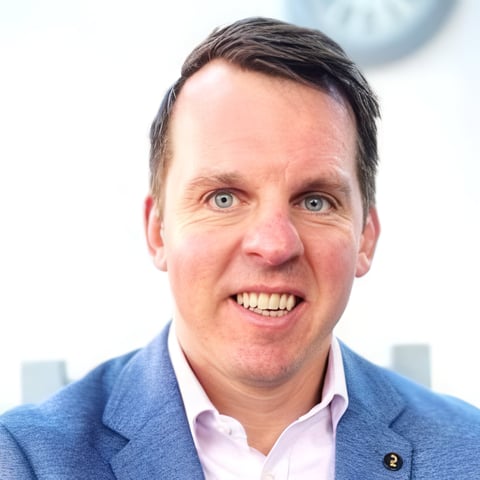 Professional headshot of a smiling man wearing a blue blazer and pink shirt