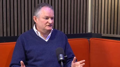 Middle-aged man in dark blue sweater seated on orange couch speaking into microphone during interview