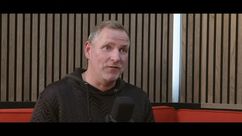 Middle-aged man in black jacket sitting indoors with microphone against striped wall background