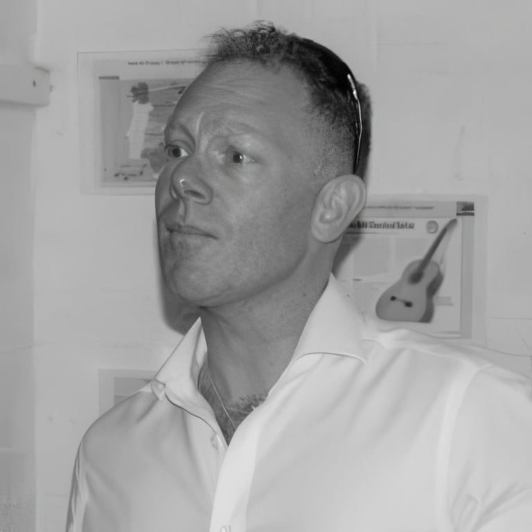 Black and white portrait of a middle-aged man in a white shirt, looking upward. Guitar images visible on the wall behind him.