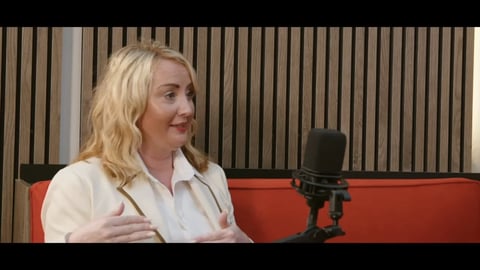Woman with blonde wavy hair in white shirt sitting at red podcast table with microphone