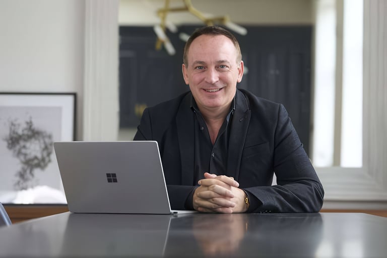 Man in dark blazer sitting at desk with silver laptop, smiling at camera in modern office setting