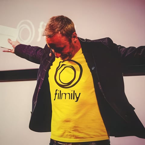 Man wearing yellow Filmily shirt and black jacket with arms outstretched against red and white background